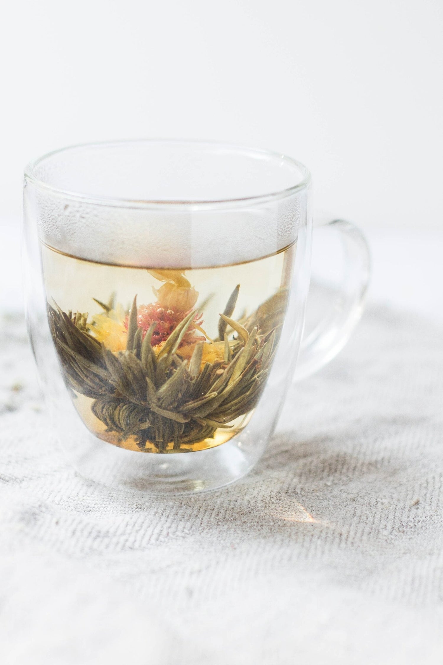 Glass mug with a blooming tea bulb steeping in pale amber water on a white linen surface