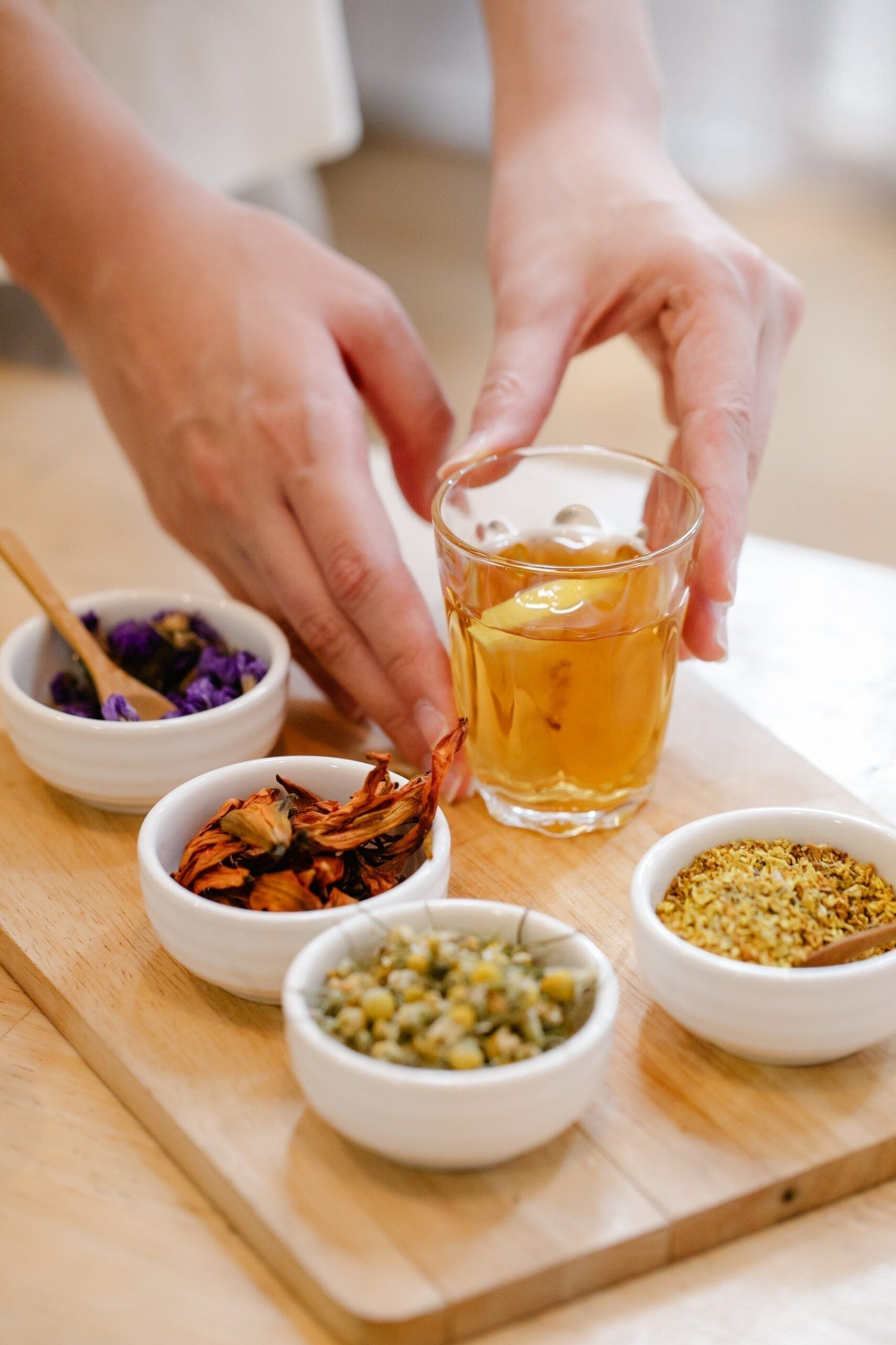Hands placing a glass of brewed herbal tea on a wooden board with small bowls of dried flowers and herbs