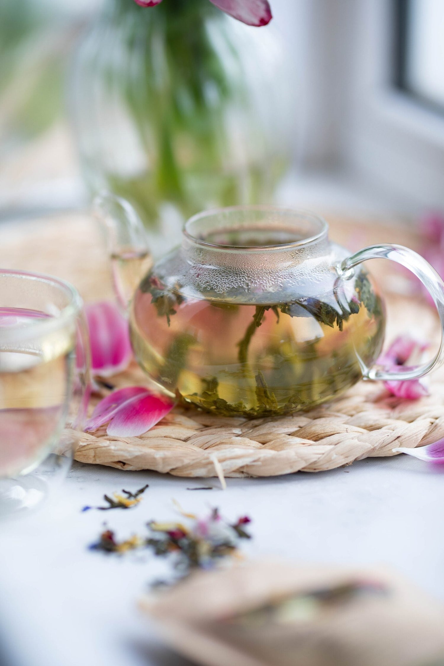 Glass teapot steeping medicinal herbs with loose tea and pink flower petals on a woven coaster by a sunlit windowsill
