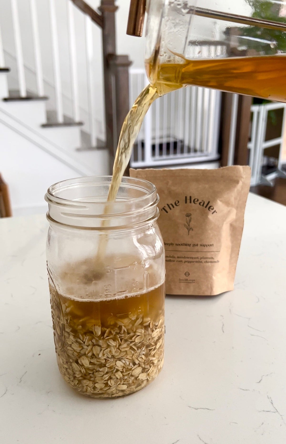 Mason jar of rolled oats being filled with warm golden liquid from a glass pitcher, kraft pouch labeled The Healer in the background