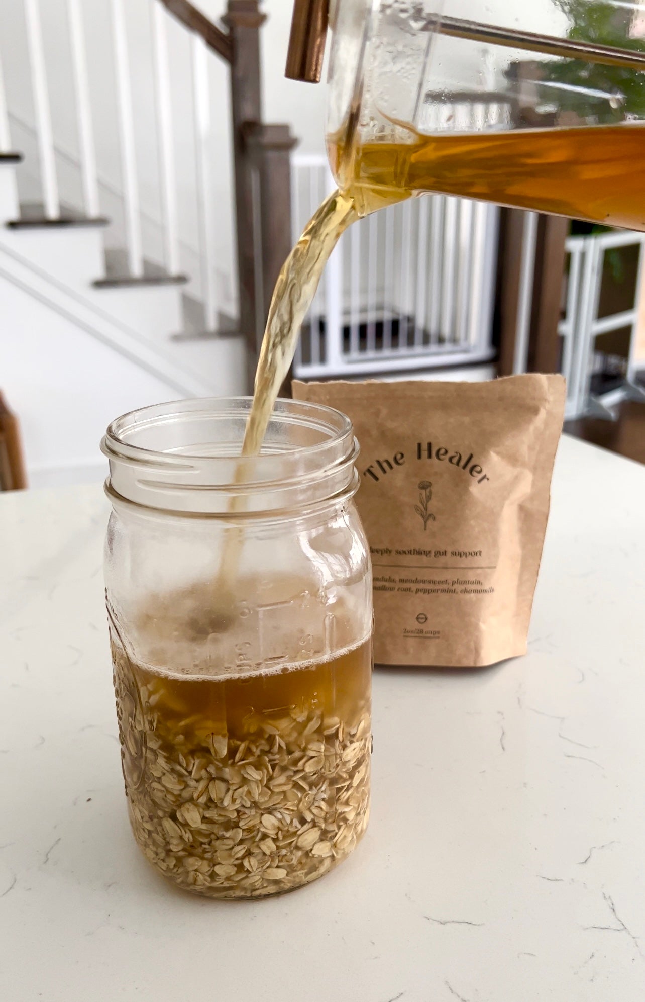 Mason jar of rolled oats being filled with warm golden liquid from a glass pitcher, kraft pouch labeled The Healer in the background