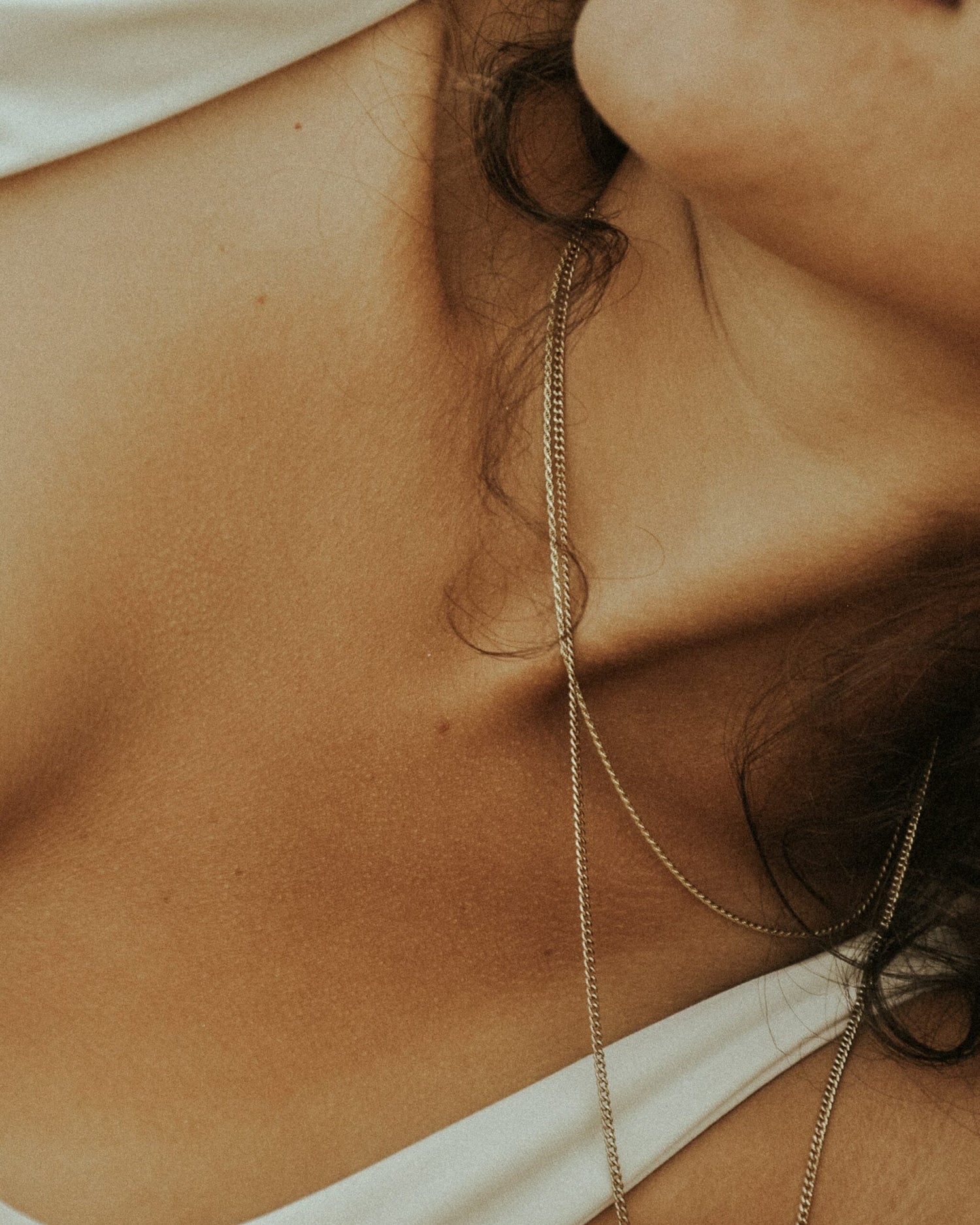 Close-up of a woman’s neck and collarbone wearing layered gold chains and a white tank top with warm lighting