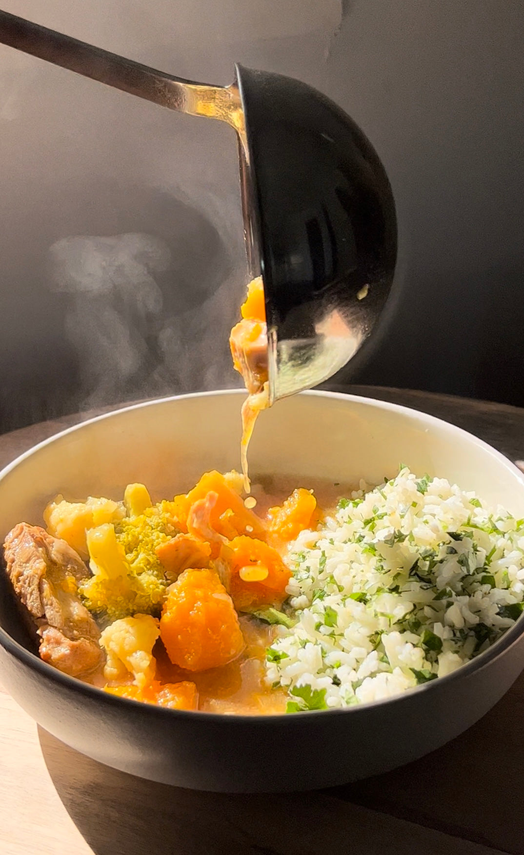 Ladle pouring steaming coconut curry chicken and vegetables into a bowl with rice and chopped cilantro