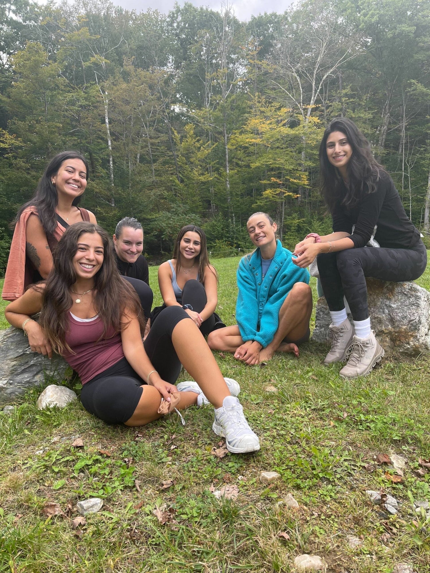 Six women sitting and smiling on grass and rocks in a forest clearing during an outdoor gathering