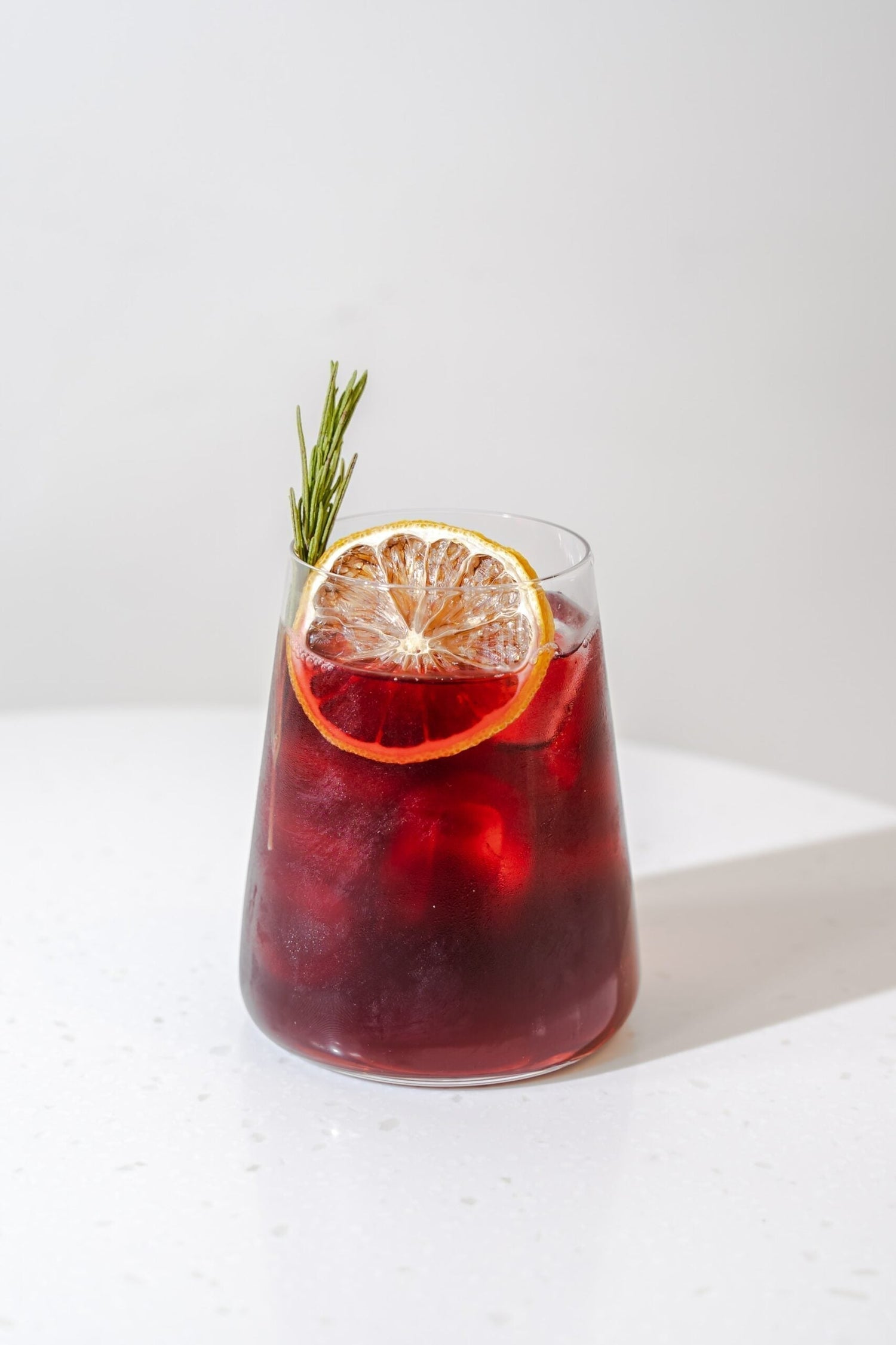 Front view of herbal hibiscus infusion in a glass showcasing a dried citrus wheel and rosemary sprig on a white countertop