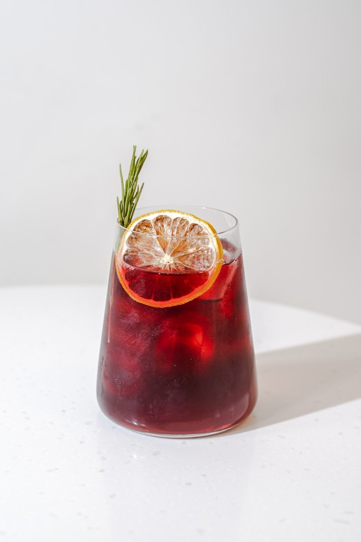 Front view of herbal hibiscus infusion in a glass showcasing a dried citrus wheel and rosemary sprig on a white countertop