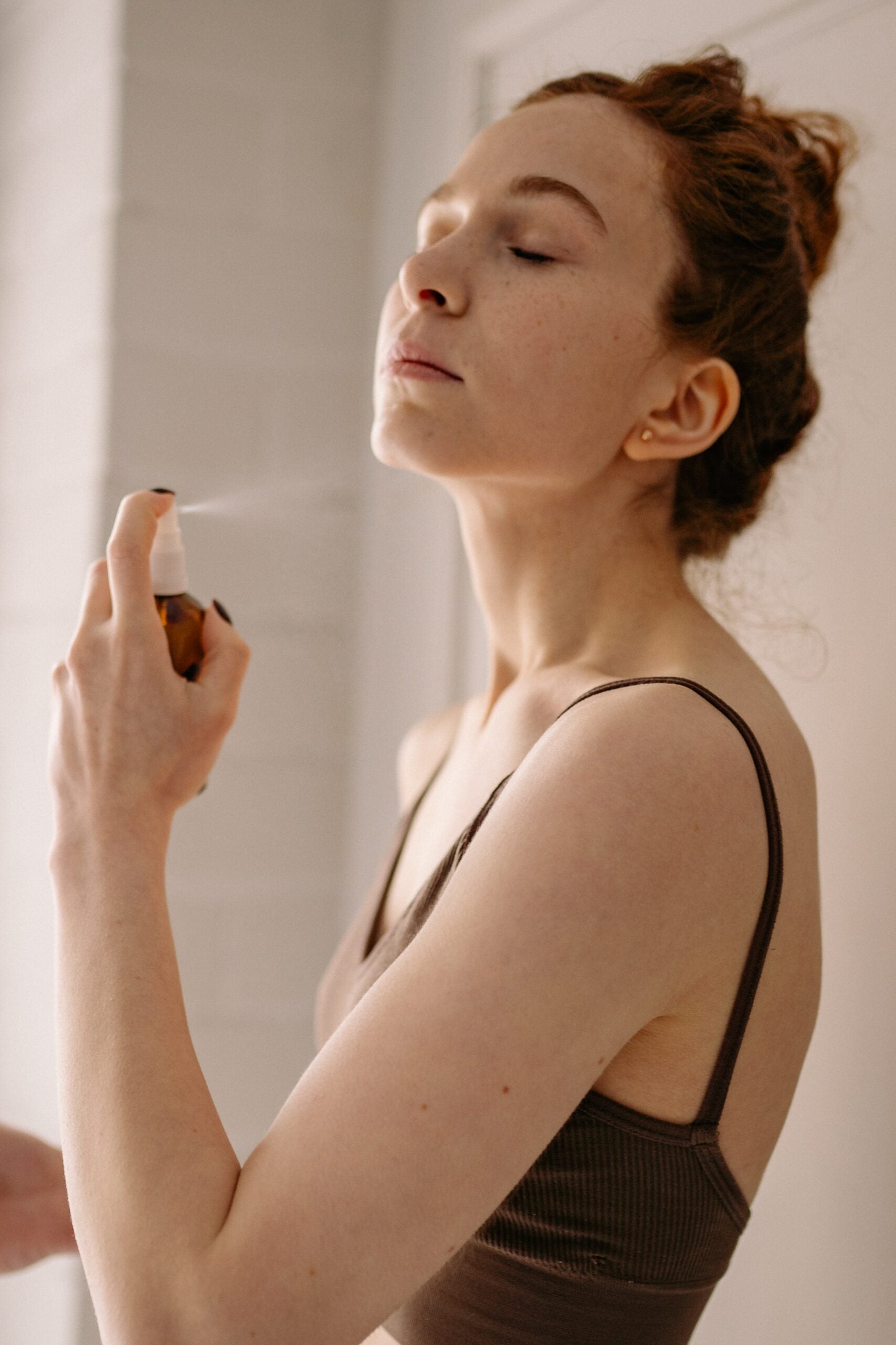 Woman with hair in a bun spraying herbal face mist onto her face in a bright bathroom