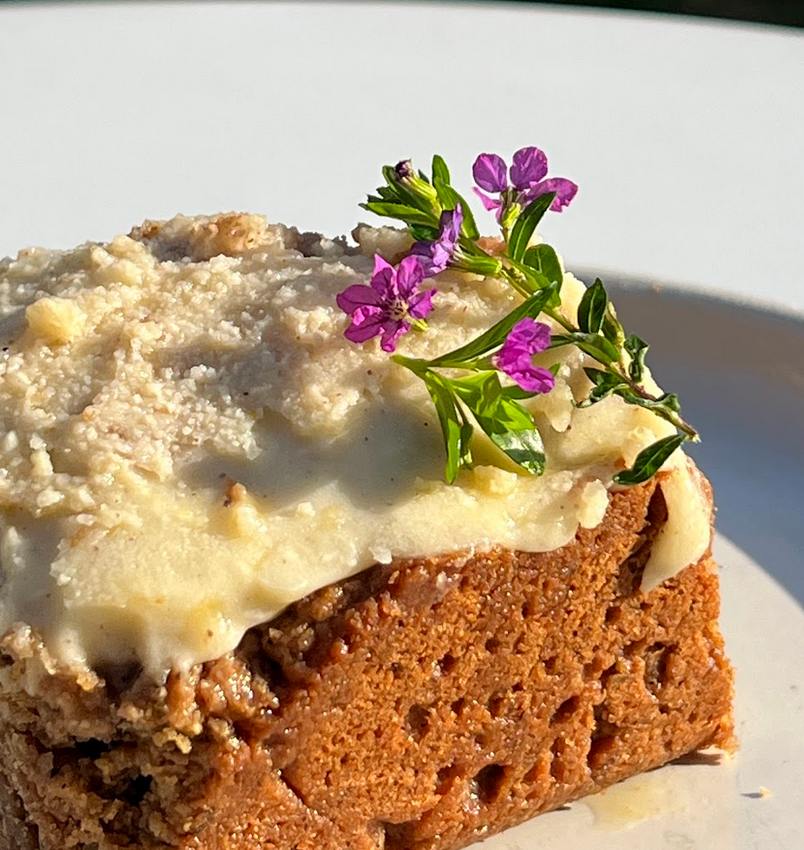 Close-up of a slice of gluten-free lavender tea cake with white frosting, crumbled topping, and purple edible flowers on a white plate