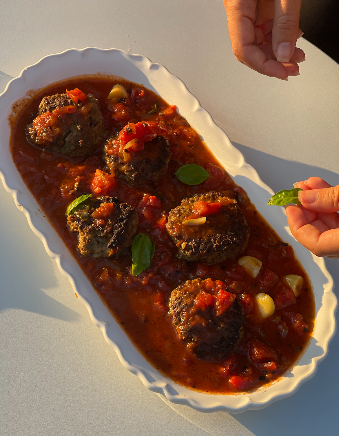 Hands placing basil on beef meatballs in chunky tomato sauce on a white oval platter, sunlight on a table