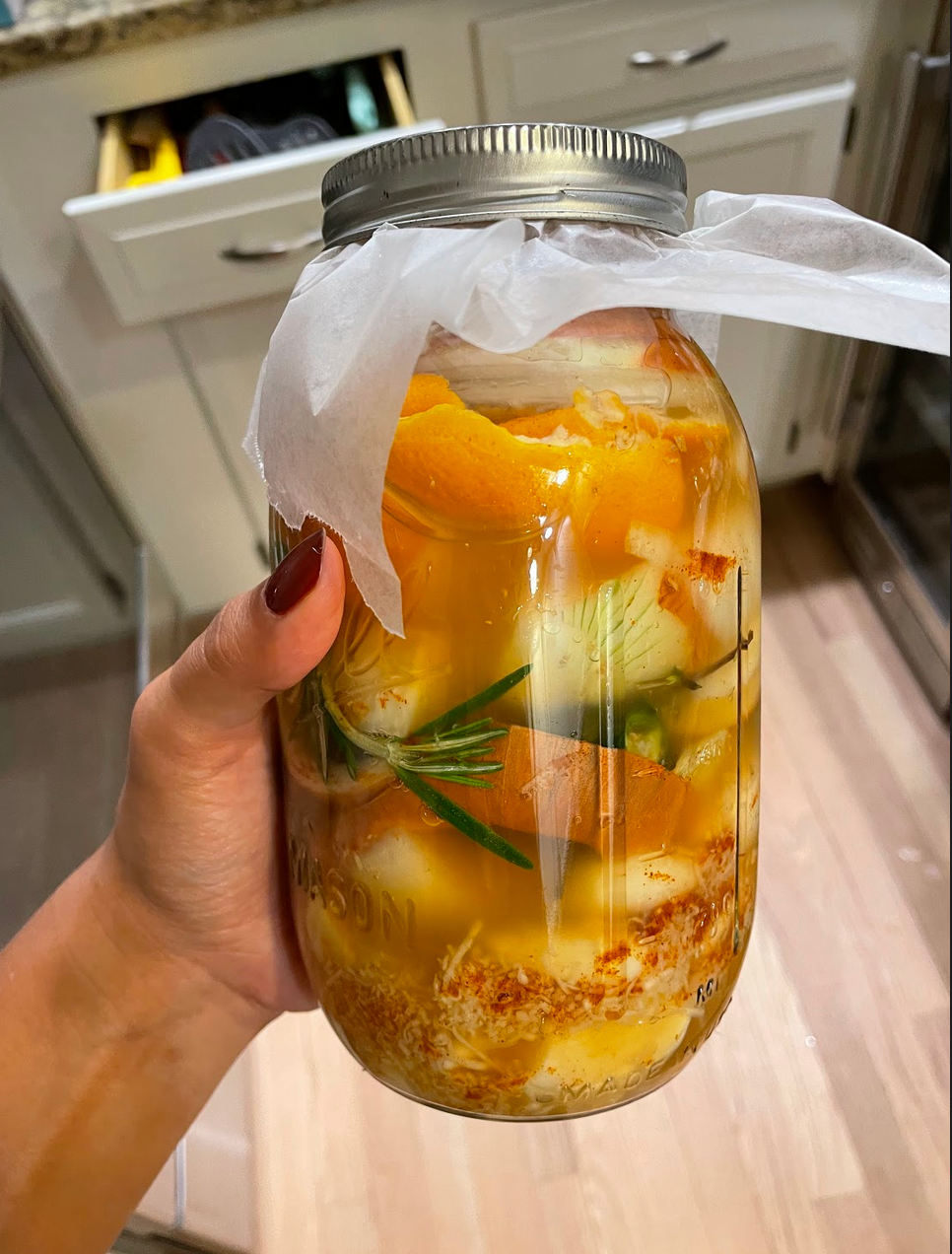 Hand holding a mason jar of fire cider with orange peels, garlic, rosemary sprig and spices in vinegar in a kitchen
