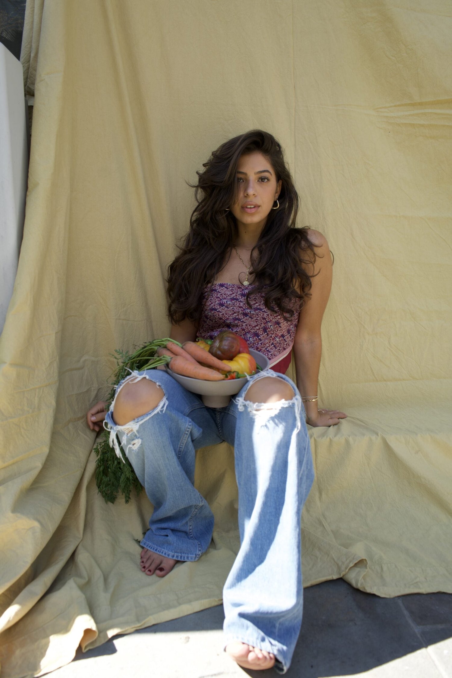 Woman sitting against a yellow backdrop holding a bowl of carrots, peaches, and greens, in a patterned top and ripped jeans