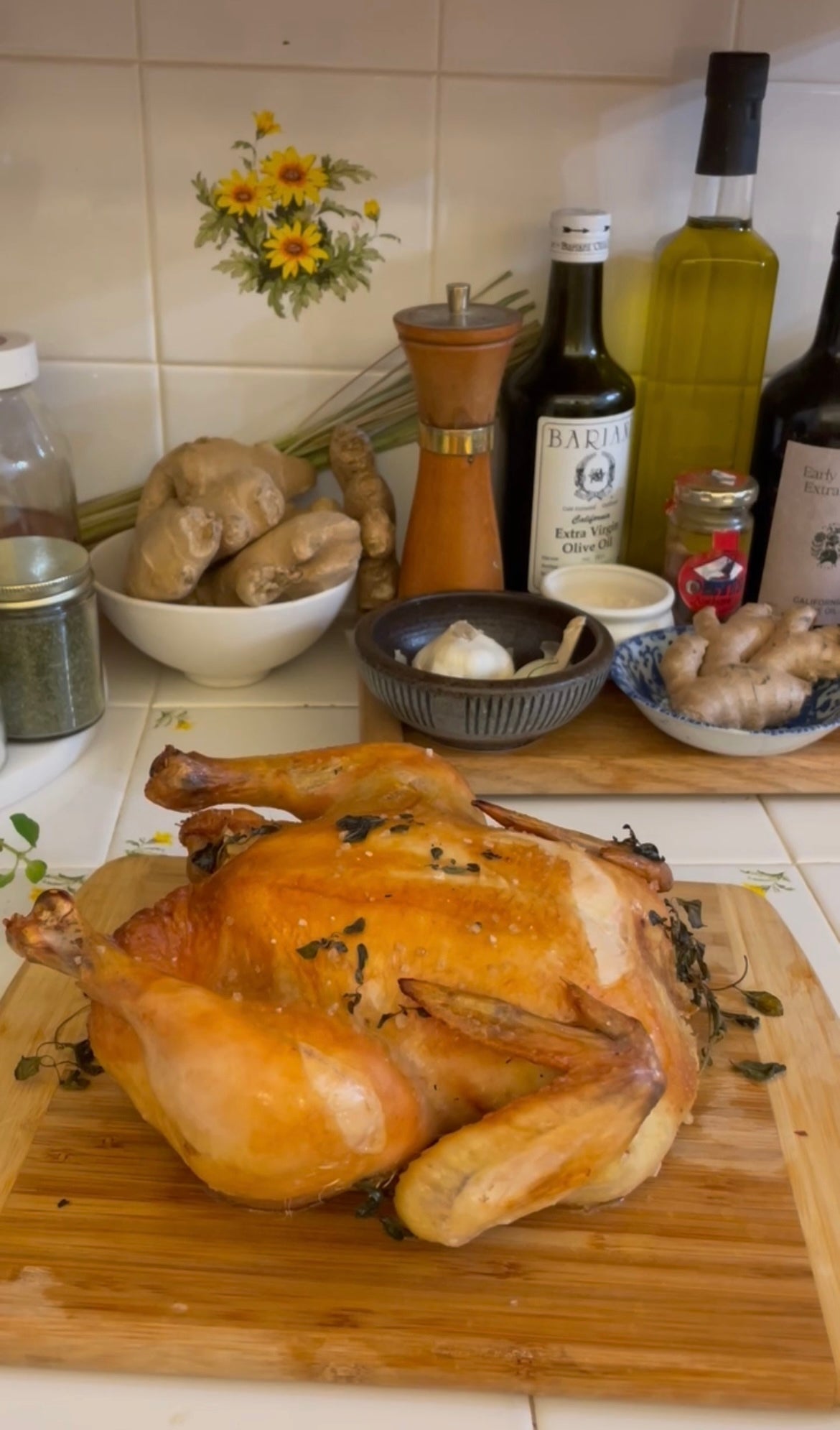 Roasted spatchcock chicken on a wooden cutting board with olive oil bottles, ginger, garlic, and spices on a tiled kitchen counter