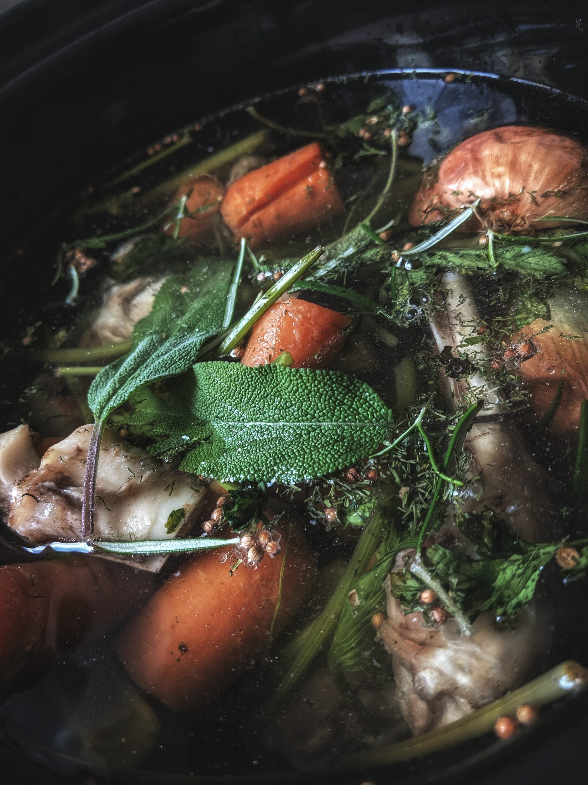 Bone broth simmering with fresh sage, carrots, onion, and herbs in a dark pot