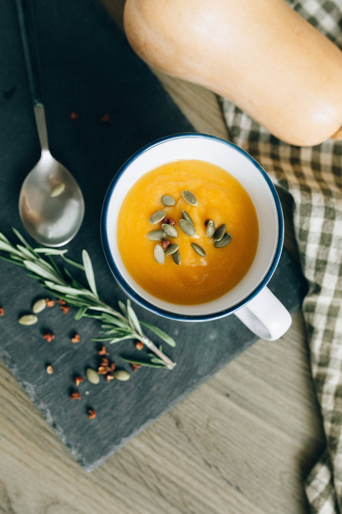 Top-down view of a mug of butternut squash soup garnished with pumpkin seeds on a slate board with a spoon and rosemary