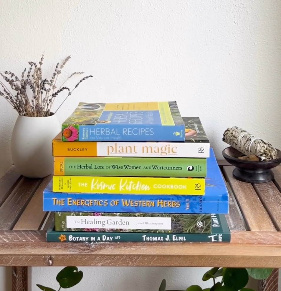 Stack of herbal books on a wooden shelf with dried lavender in a white vase to the left and a sage bundle on a dish to the right against a pale wall