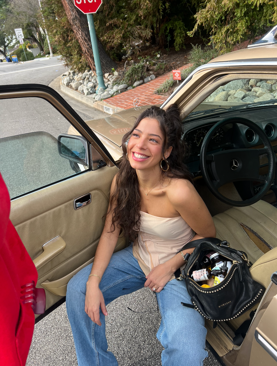 Smiling woman sitting in the open door of a Mercedes, purse with supplement bottles on her lap, parked on a street