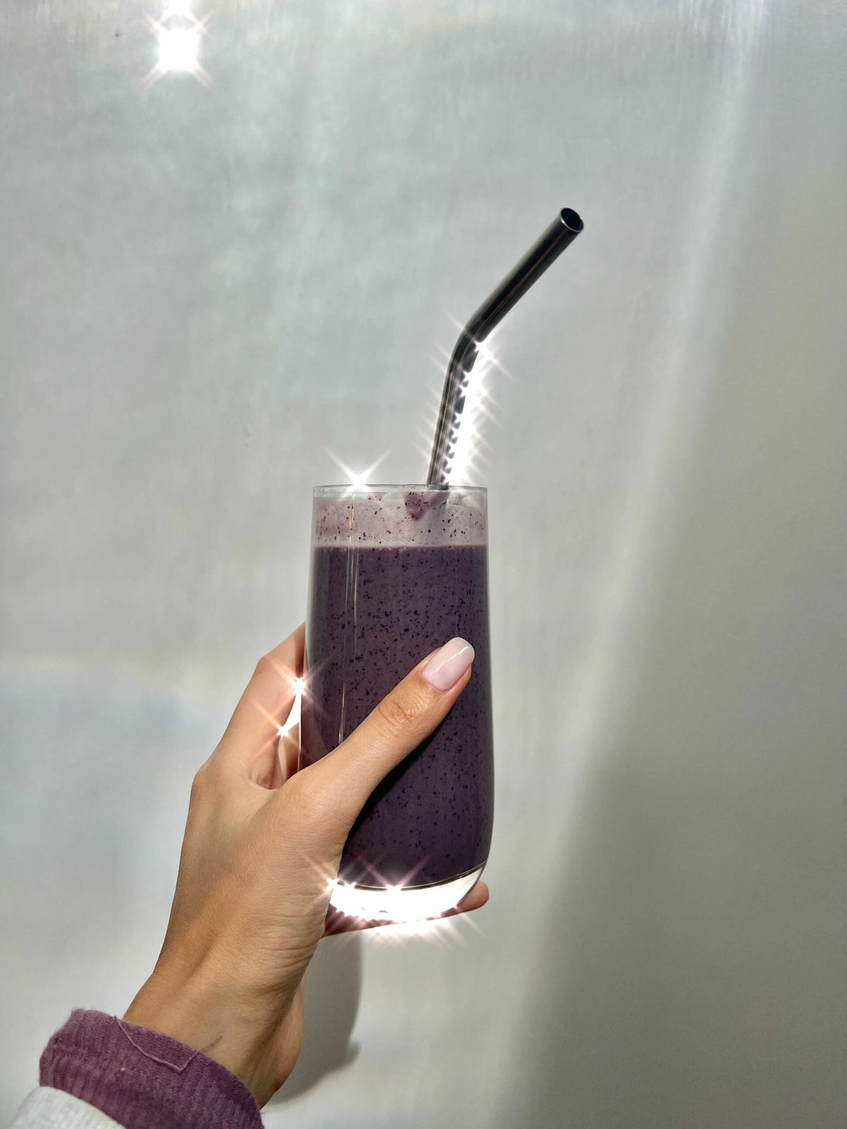 Hand holding a tall glass of purple blueberry tahini protein smoothie with a bent stainless steel straw against a soft gray background