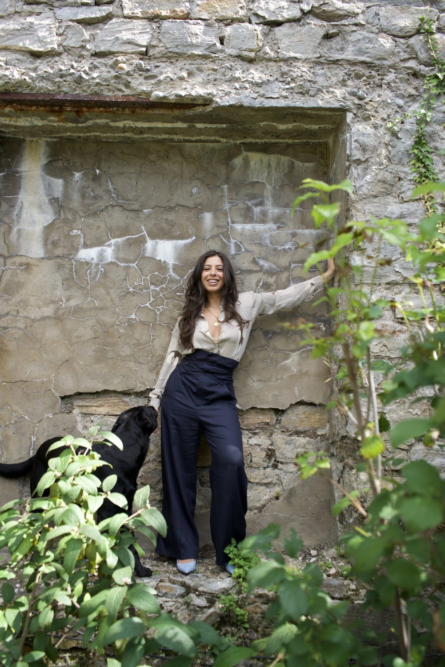 Woman smiling and leaning against a cracked stone wall while petting a black dog in a leafy outdoor setting