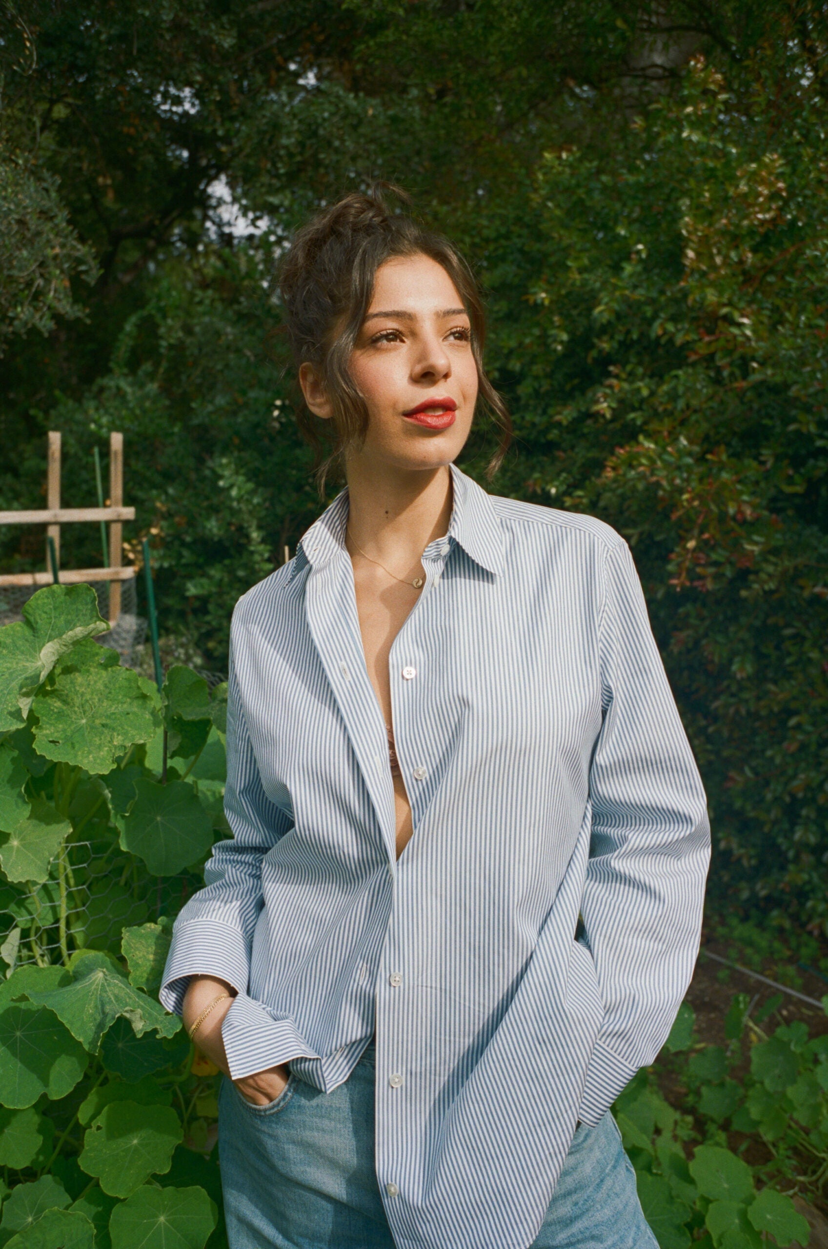 Woman standing in a sunlit garden looking off to the side, wearing a blue-and-white striped shirt with hands in pockets