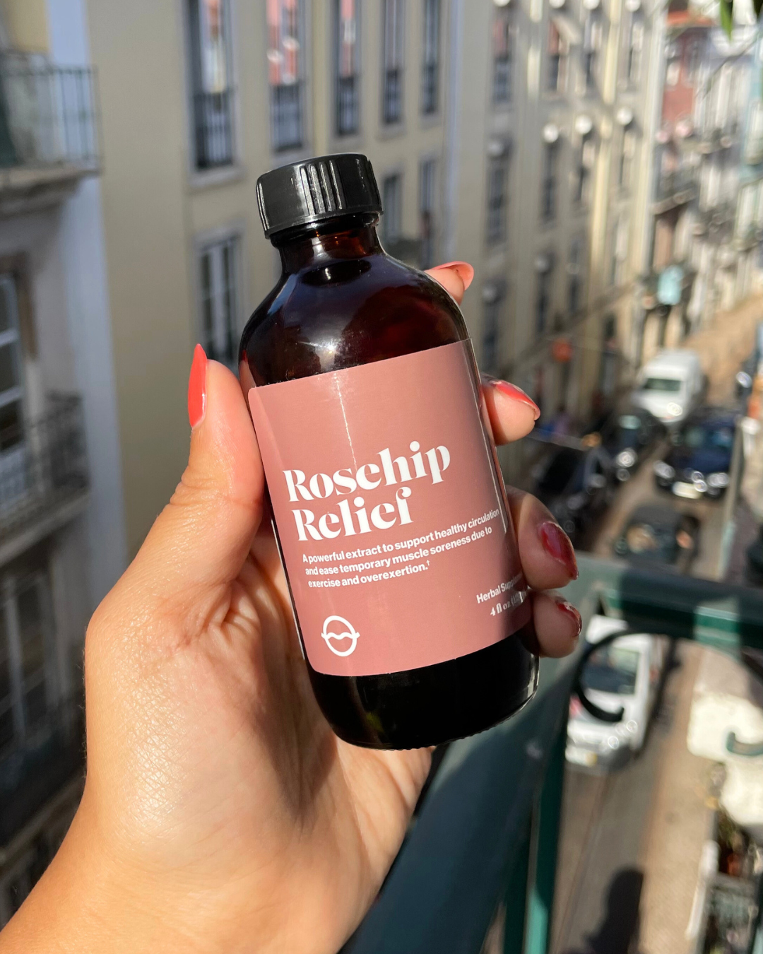 Front view of Rosehip Relief amber glass bottle showcasing a pink label reading ‘Rosehip Relief’ held in hand over a sunlit city street