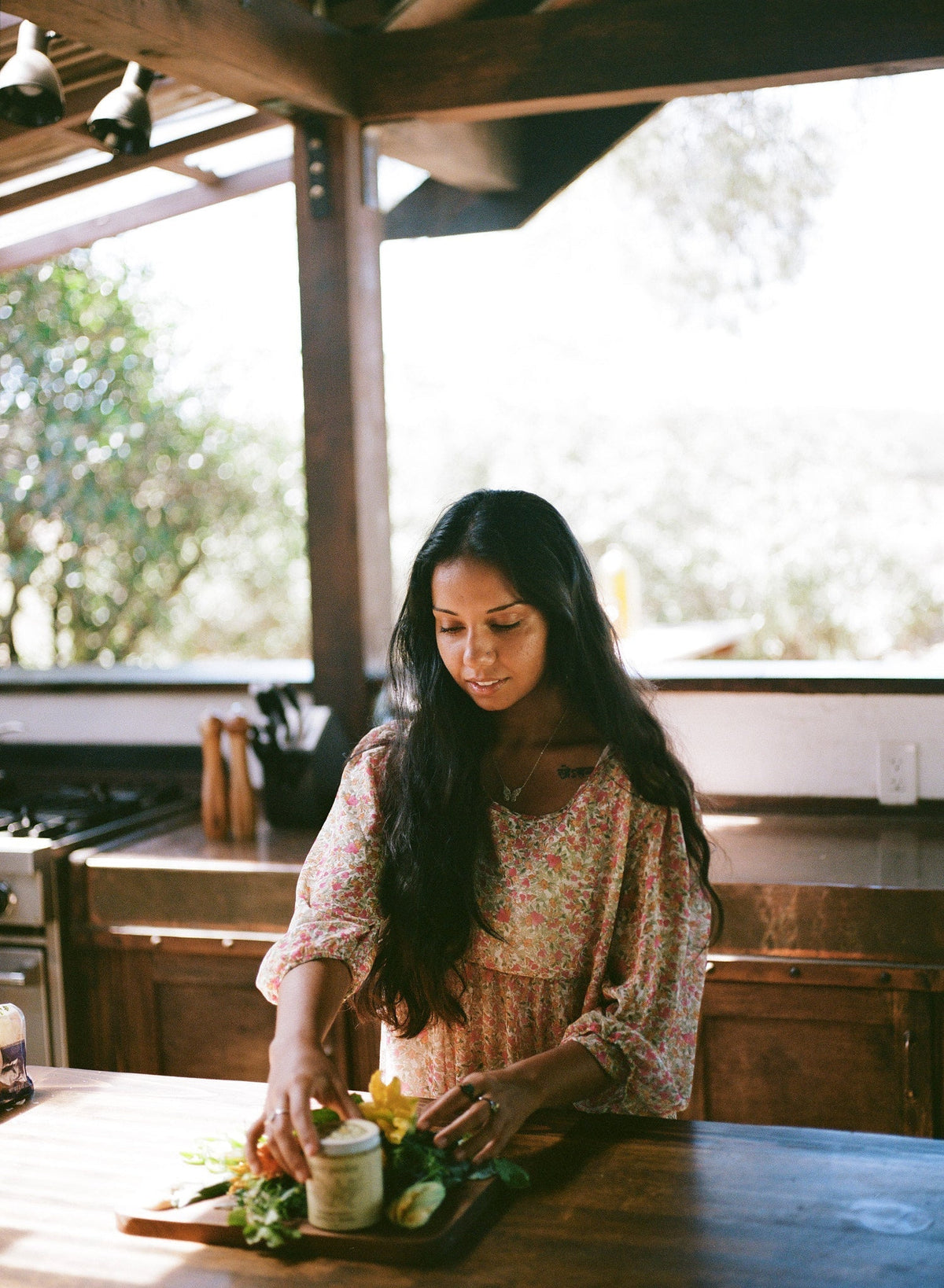 Woman arranging dandelion greens and herbs with a jar of pesto on a wooden board in a sunlit rustic kitchen