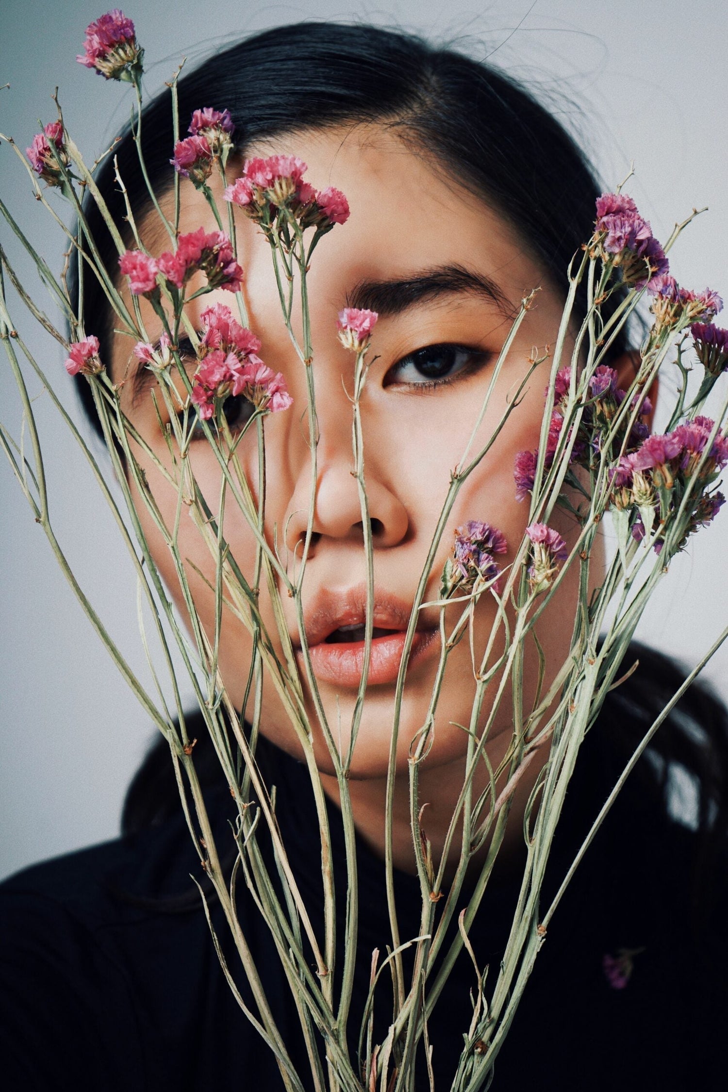 Close-up portrait of a young woman partially hidden behind stems of dried pink flowers against a neutral background
