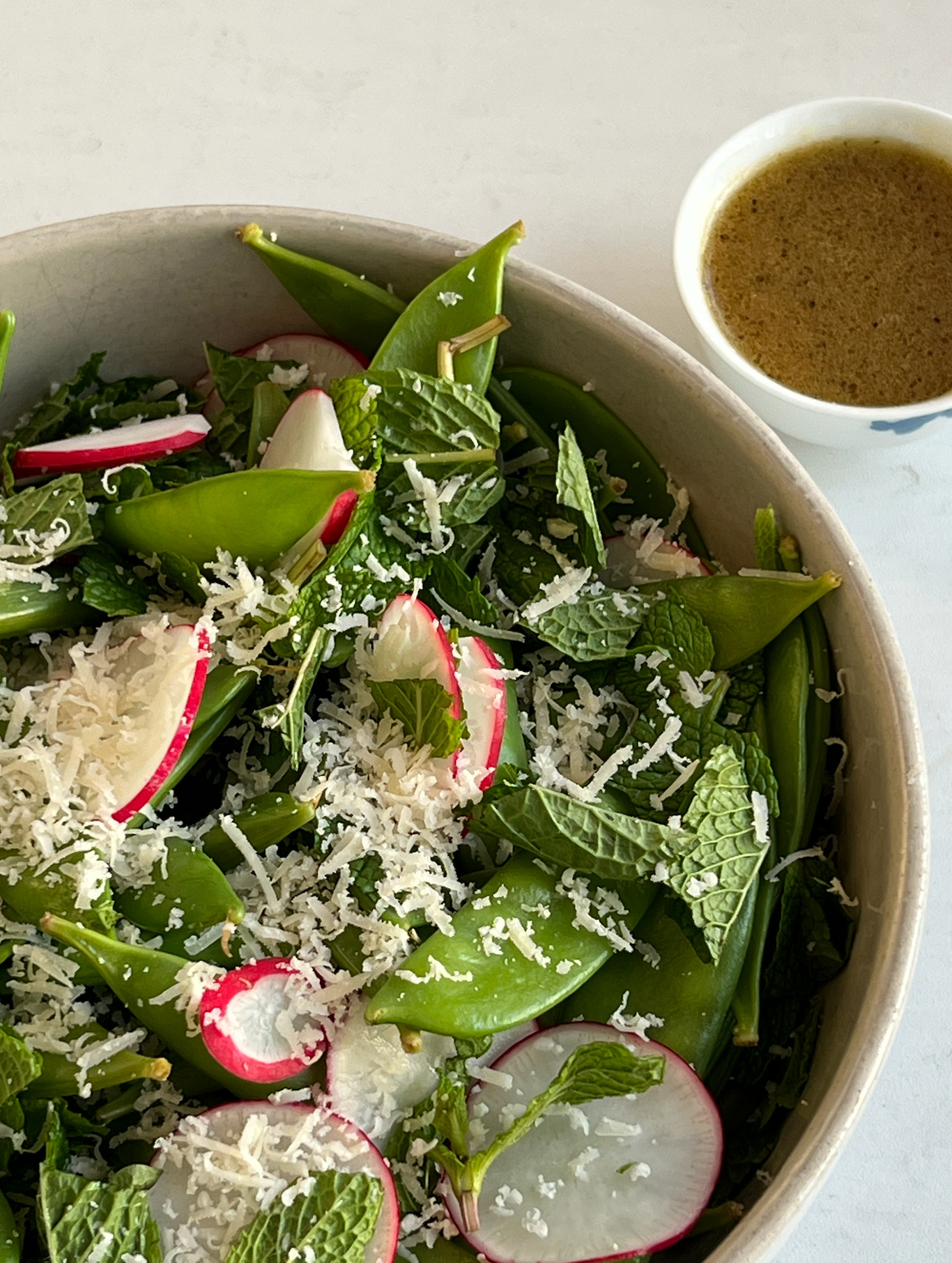 Warm snap pea salad in a ceramic bowl with sliced radishes, fresh mint, grated cheese, and a small bowl of vinaigrette on a white table