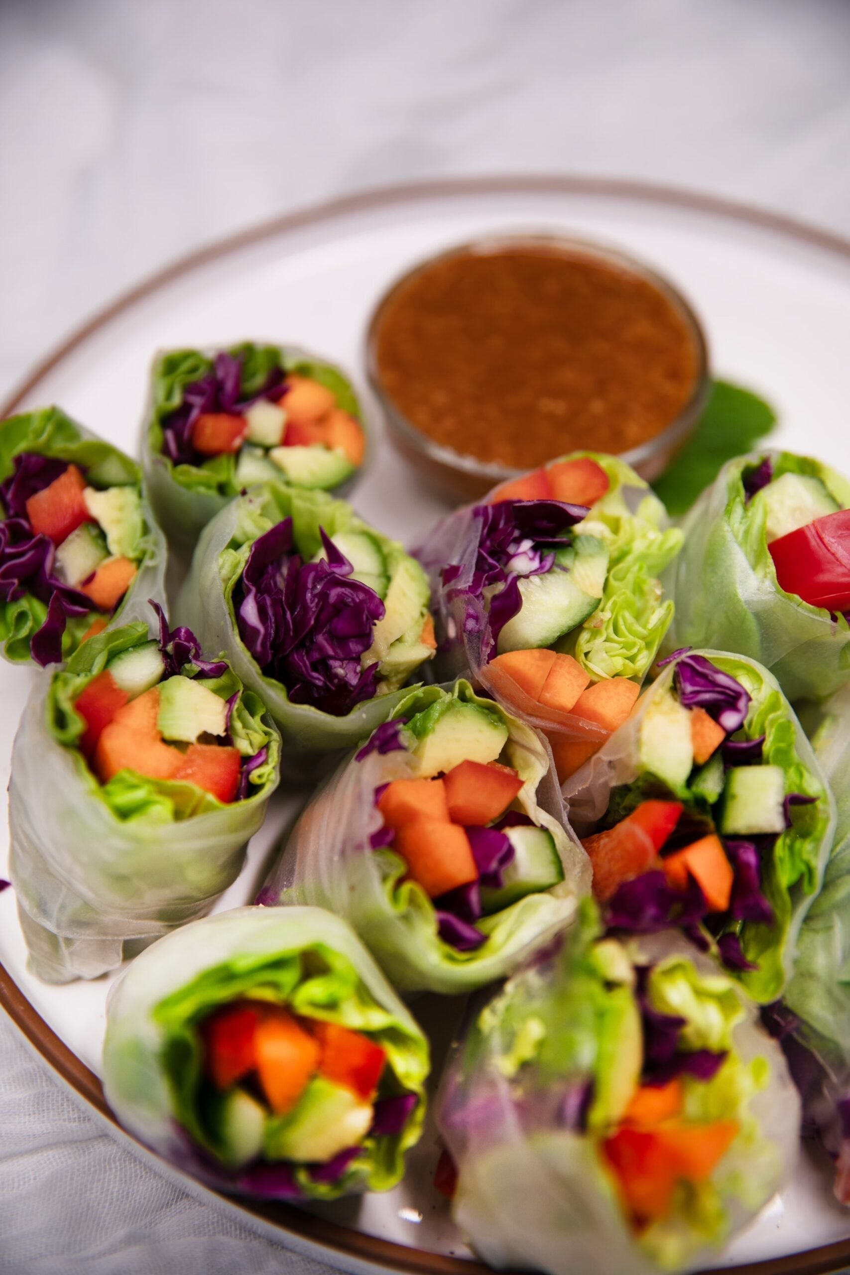 Rainbow spring rolls on a white plate with purple cabbage, cucumber, carrot, red pepper, and peanut dipping sauce