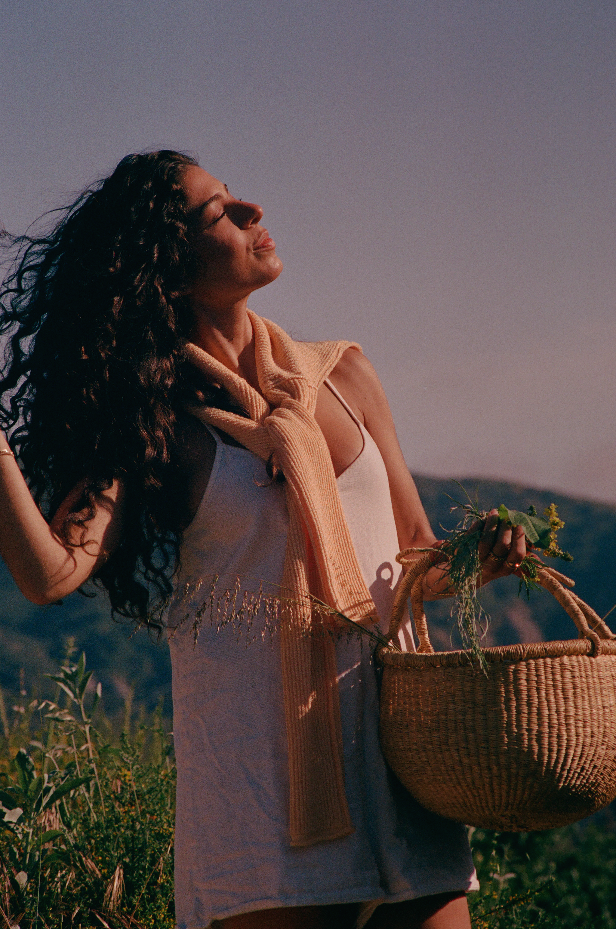 Woman with curly hair tilting her face toward the sun while holding a woven basket of wild plants in a grassy hillside