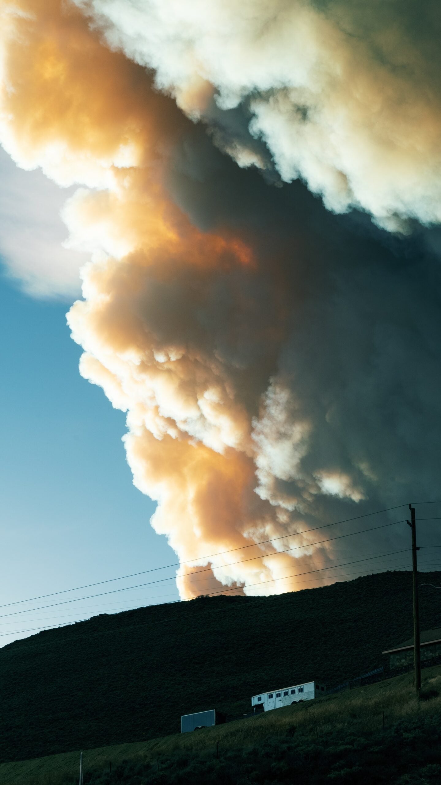 Plume of wildfire smoke billowing above a hillside, clouds lit orange by sunlight