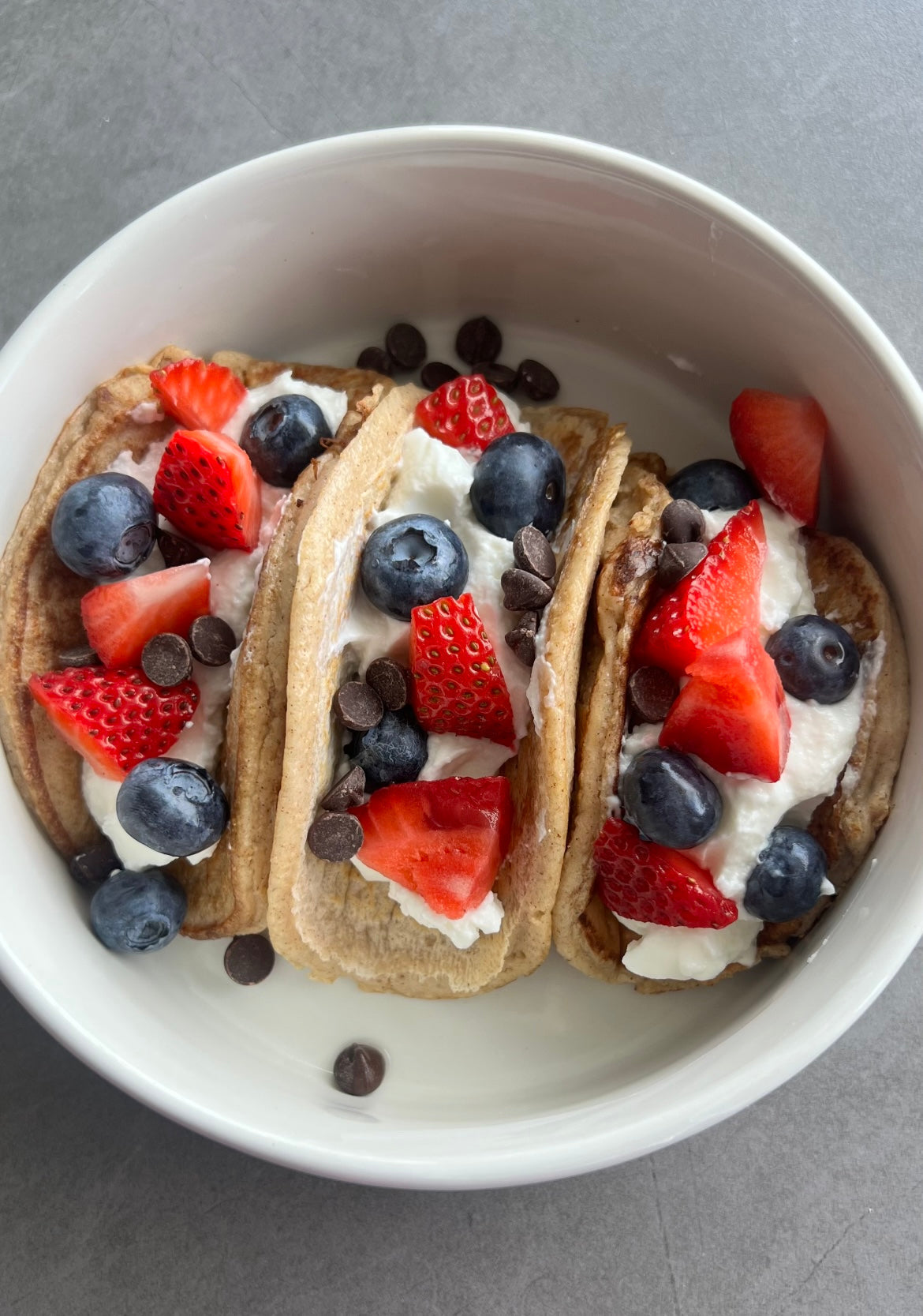 Three folded pancakes with white creamy filling, strawberries, blueberries, and chocolate chips in a white bowl
