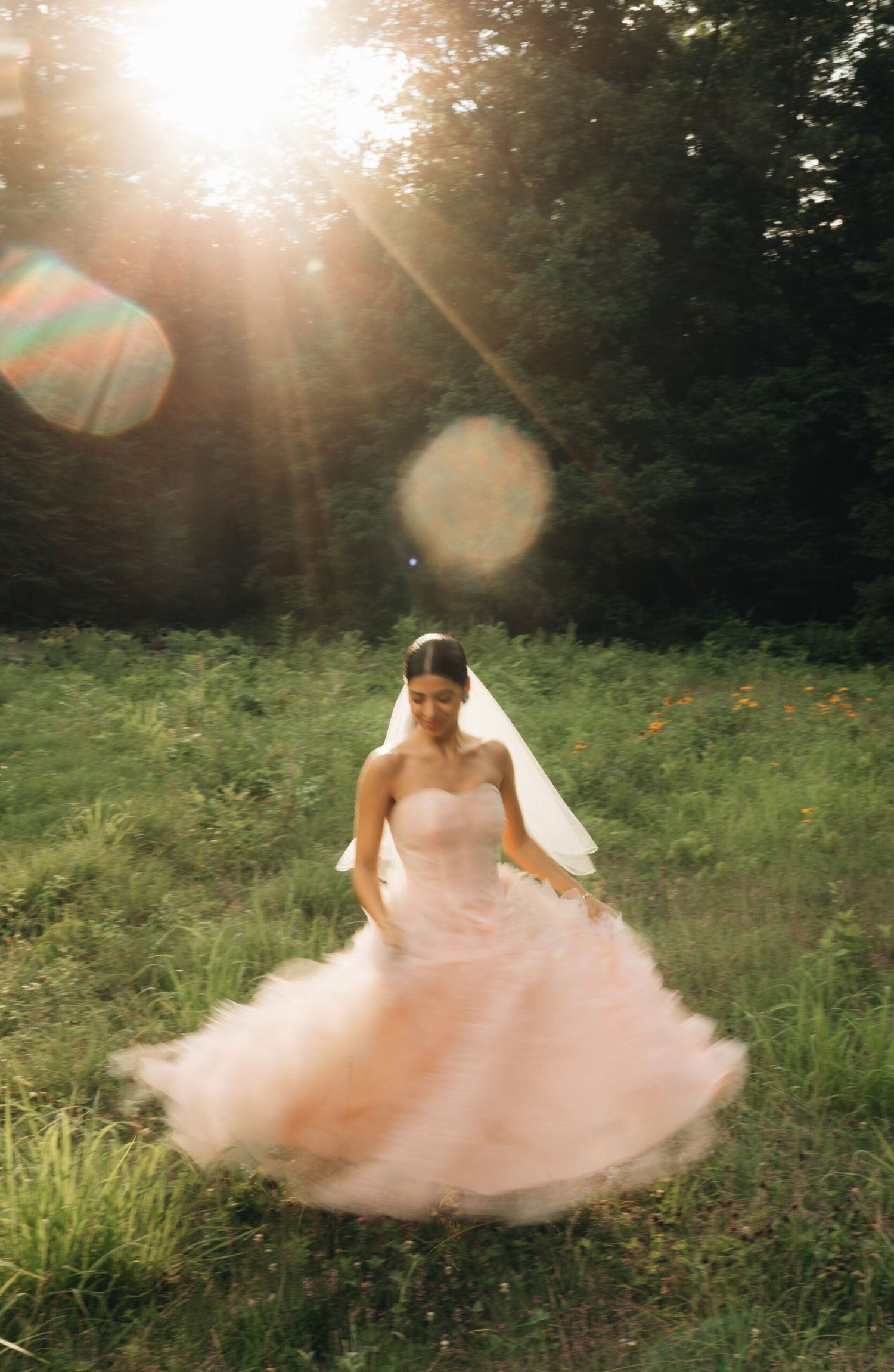 Bride spinning in a strapless blush vintage wedding gown and veil in a sunlit meadow with lens flare