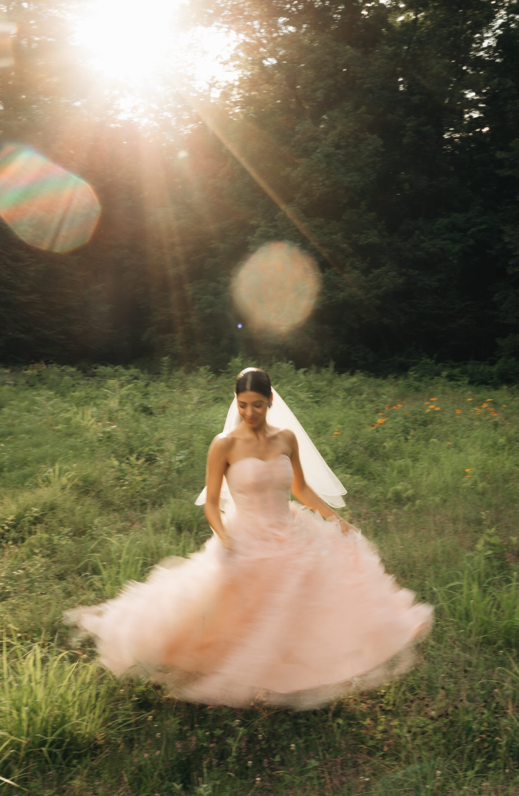 Bride spinning in a strapless blush vintage wedding gown and veil in a sunlit meadow with lens flare
