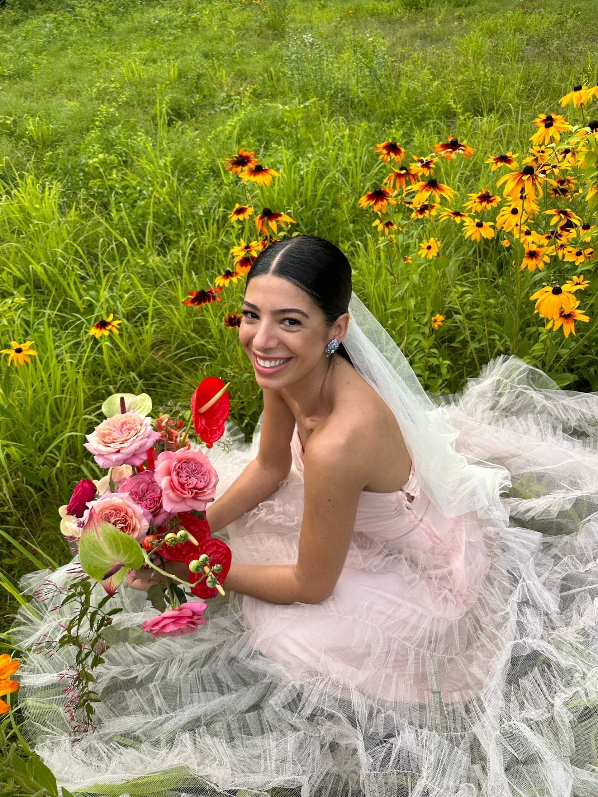Bride in a pink vintage wedding gown smiling and holding a pink and red bouquet while seated in a grassy field of yellow wildflowers