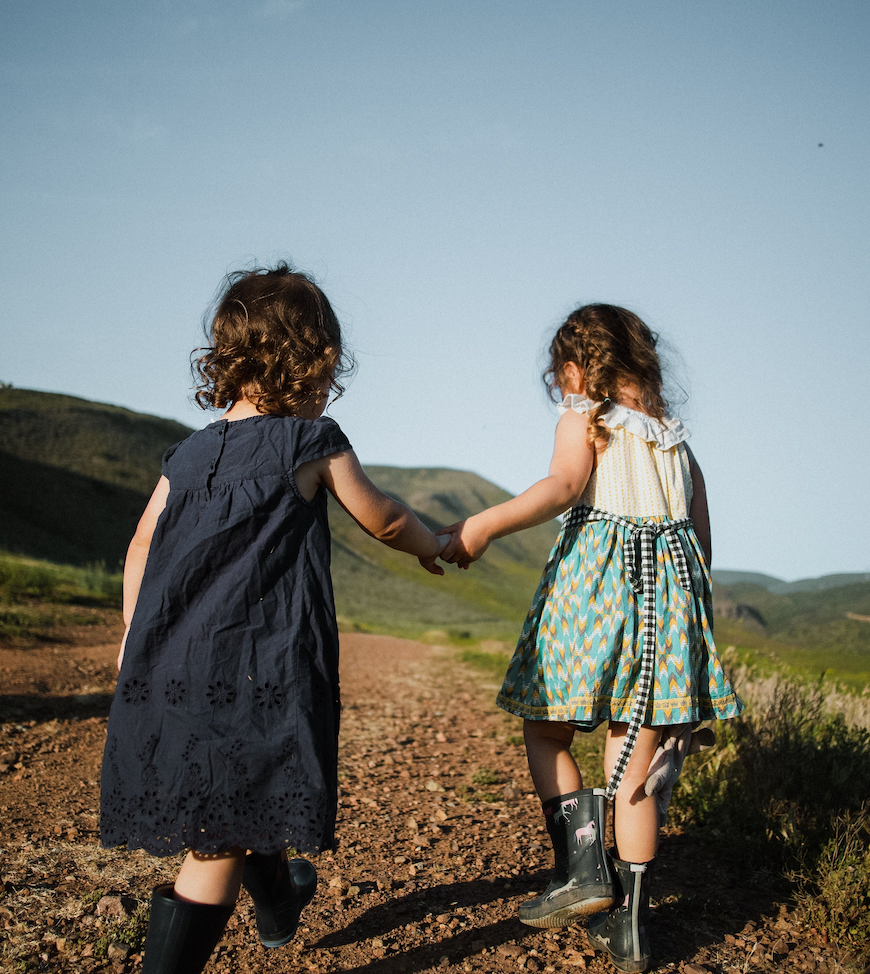 Two young girls holding hands as they walk on a sunlit dirt path with grassy hills in the background