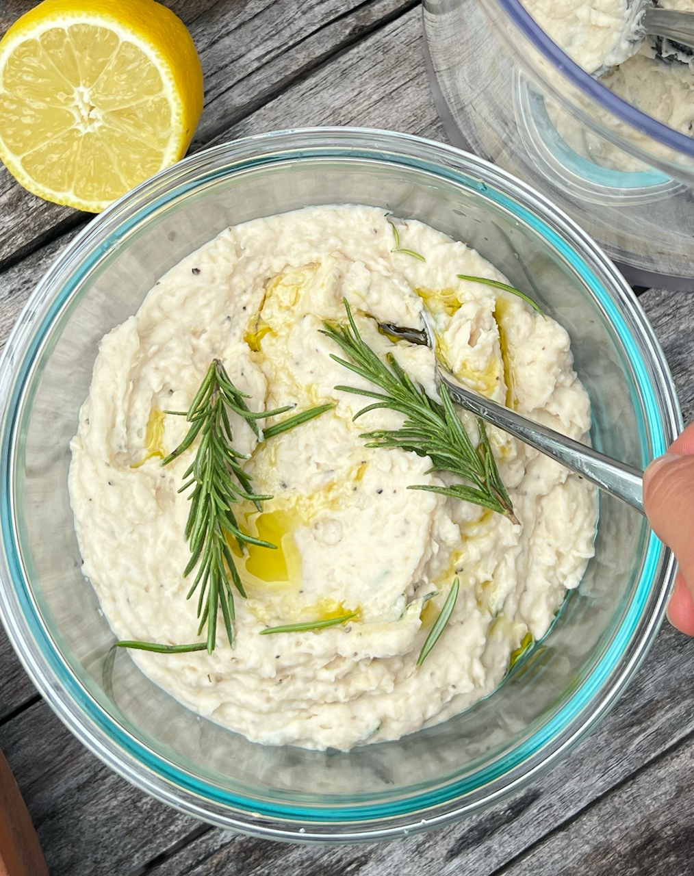Top view of high-fiber herby bean dip in a glass bowl showing creamy texture, rosemary sprigs and olive oil drizzle on wood