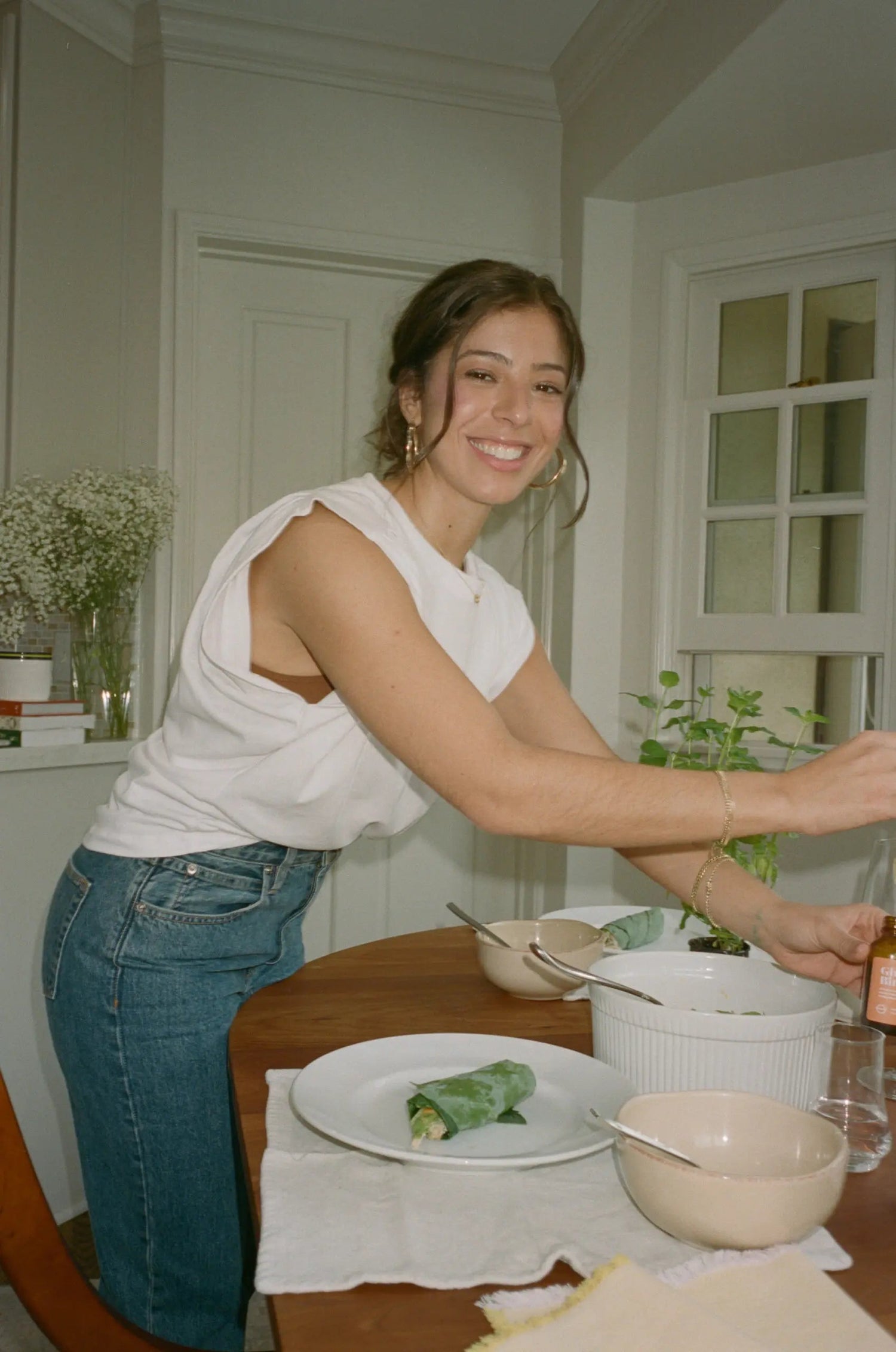 Smiling woman leaning over a dining table preparing a leafy wrap in a bright home kitchen