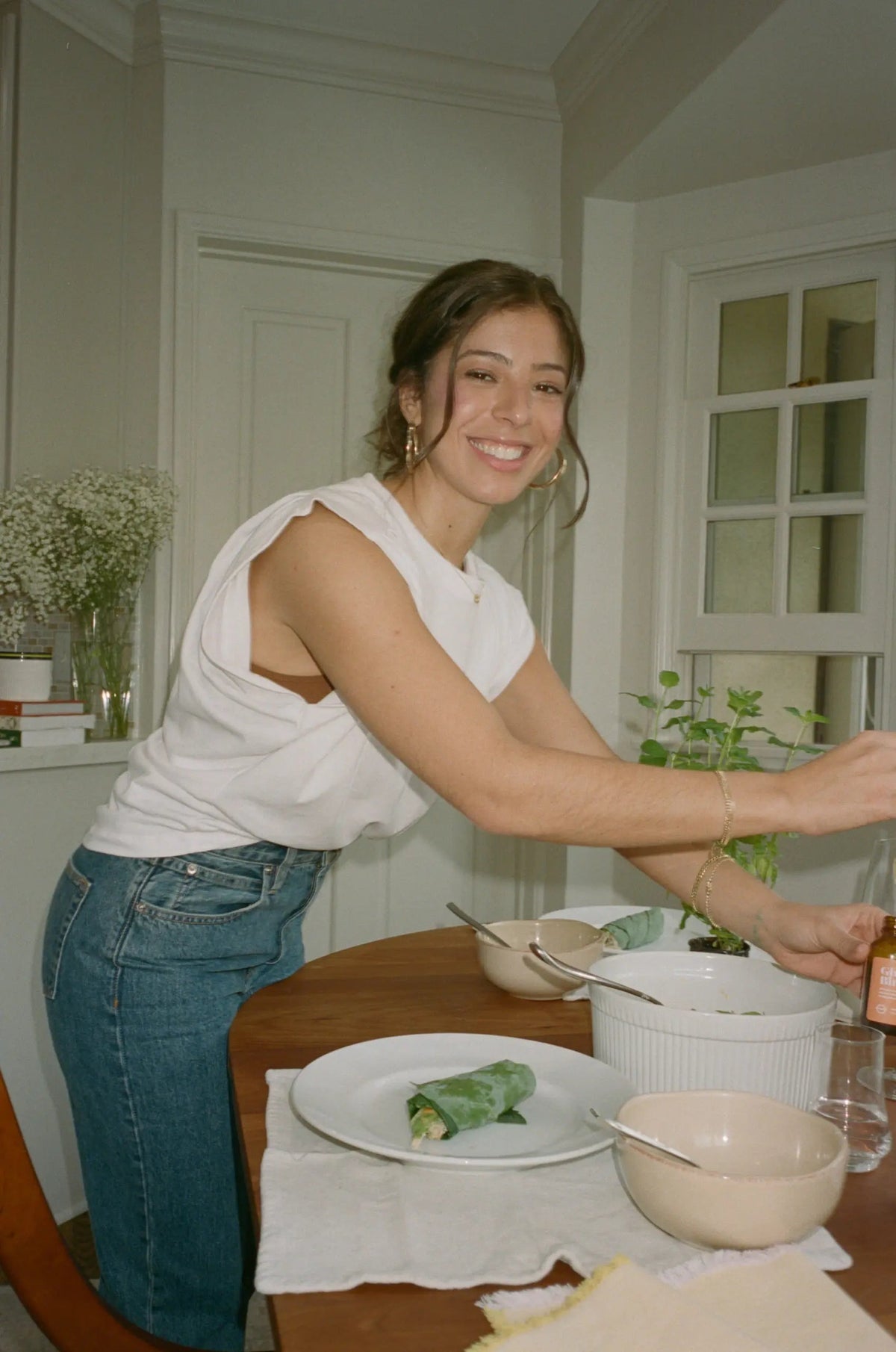 Smiling woman leaning over a dining table preparing a leafy wrap in a bright home kitchen