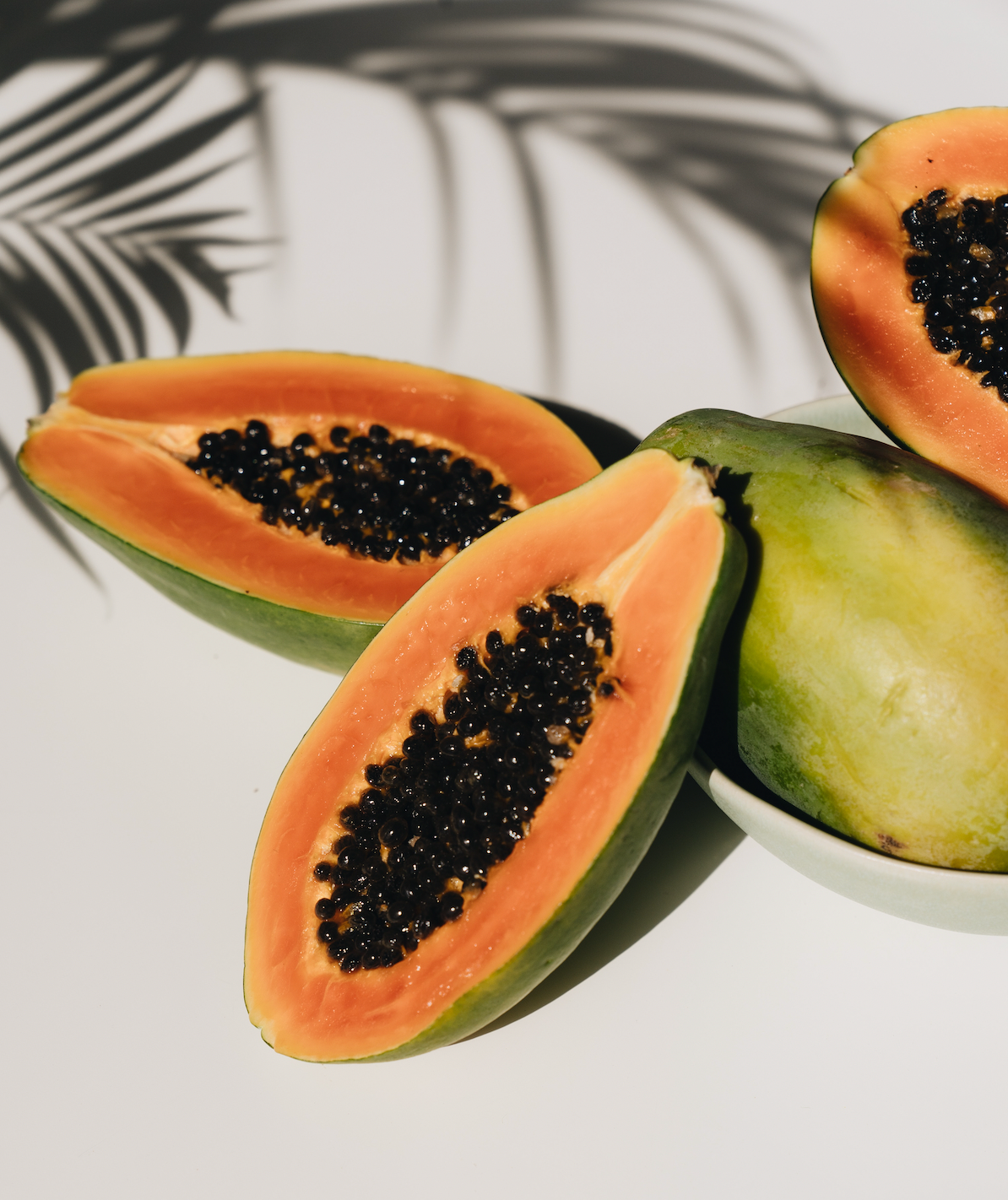 Halved ripe papayas arranged on a white surface showing orange flesh and black seeds with a palm-leaf shadow in the background