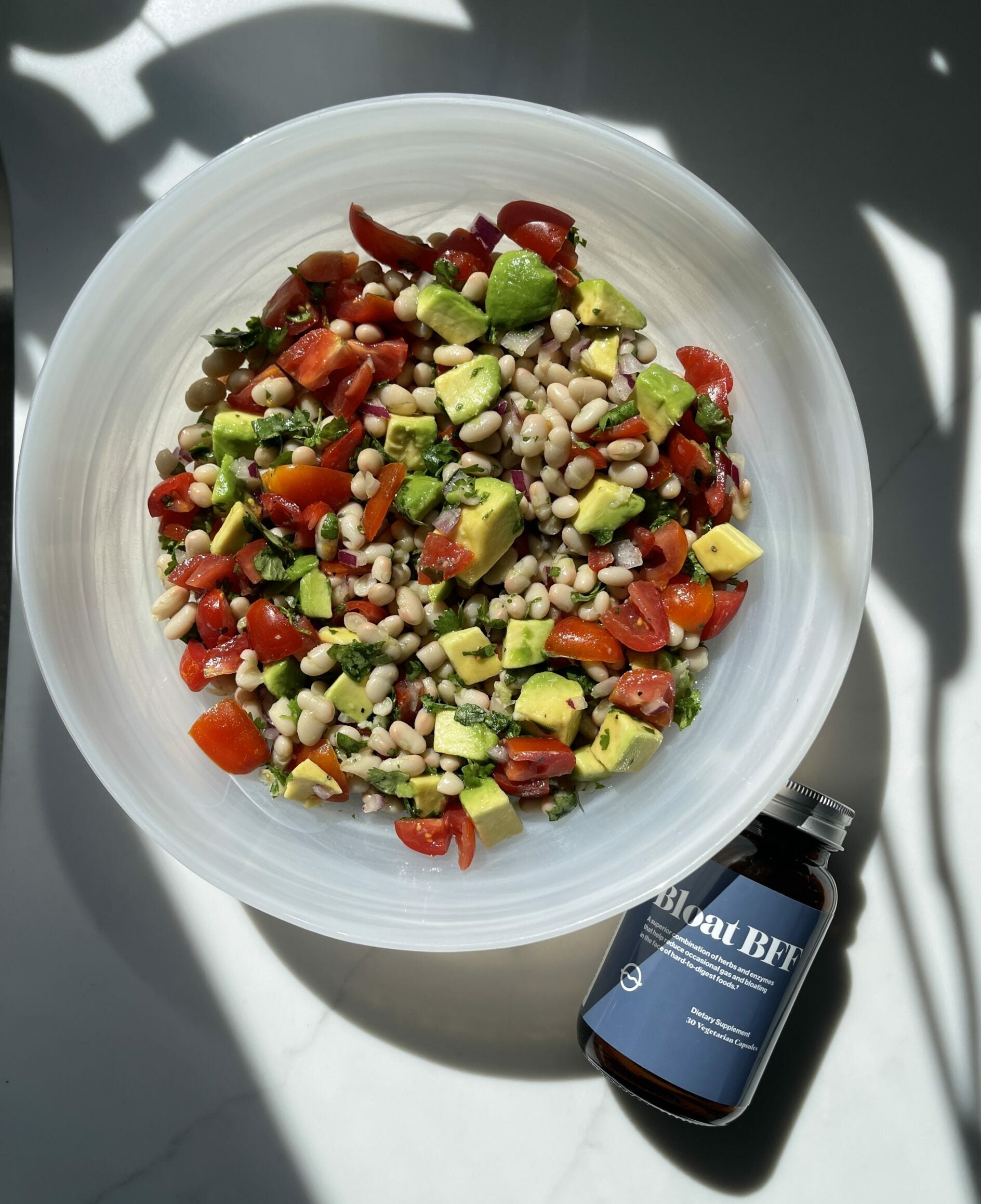White bean salad with avocado, cherry tomatoes, red onion, and herbs in a bowl beside a Bloat BFF bottle on sunlit marble