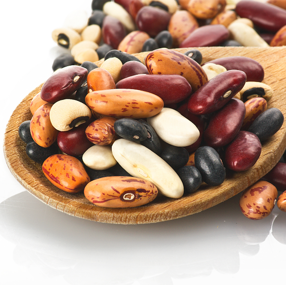 Close-up of mixed dried beans in a wooden spoon showcasing varied colors, sizes, and patterns on a white background