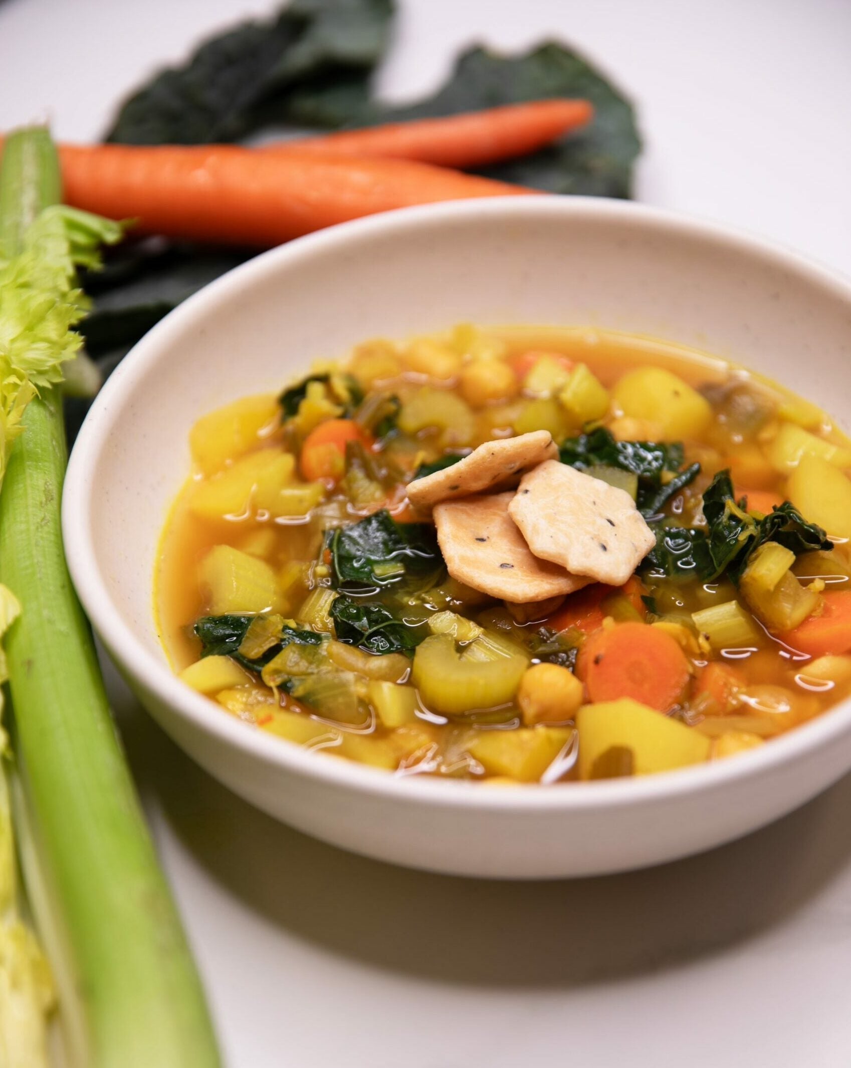 Bowl of ginger turmeric vegetable soup with carrots, celery, kale, and chickpeas, topped with crackers, with whole carrots and celery beside the bowl