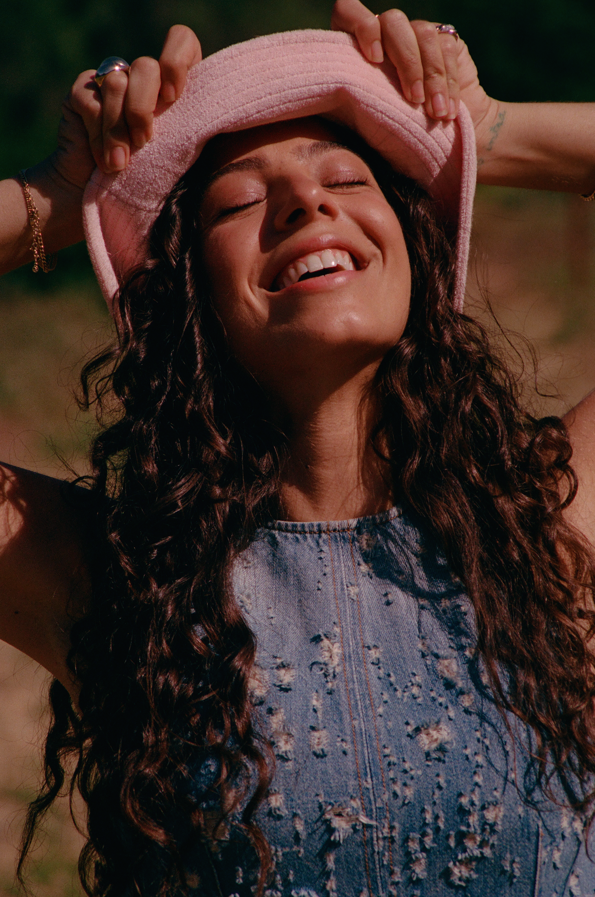 Woman with long curly hair smiling with eyes closed while holding a pink bucket hat outdoors in warm sunlight