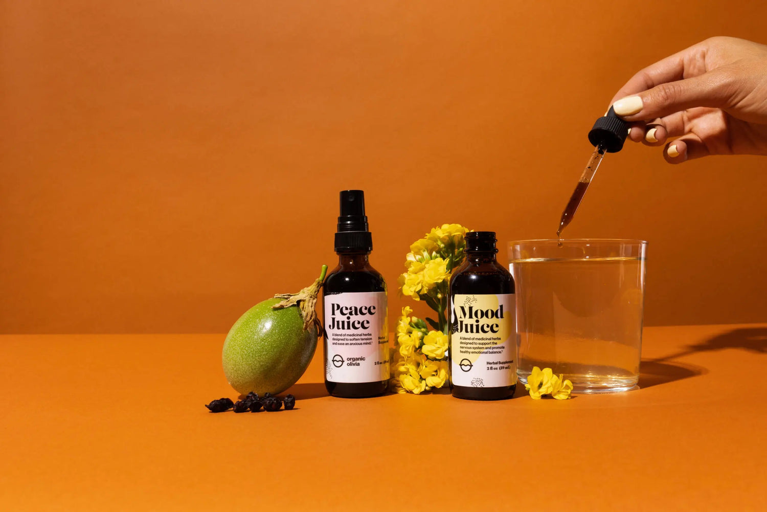 Front view of two amber bottles labeled Peace Juice and Mood Juice with dropper, fruit, yellow flowers, and a glass on an orange background