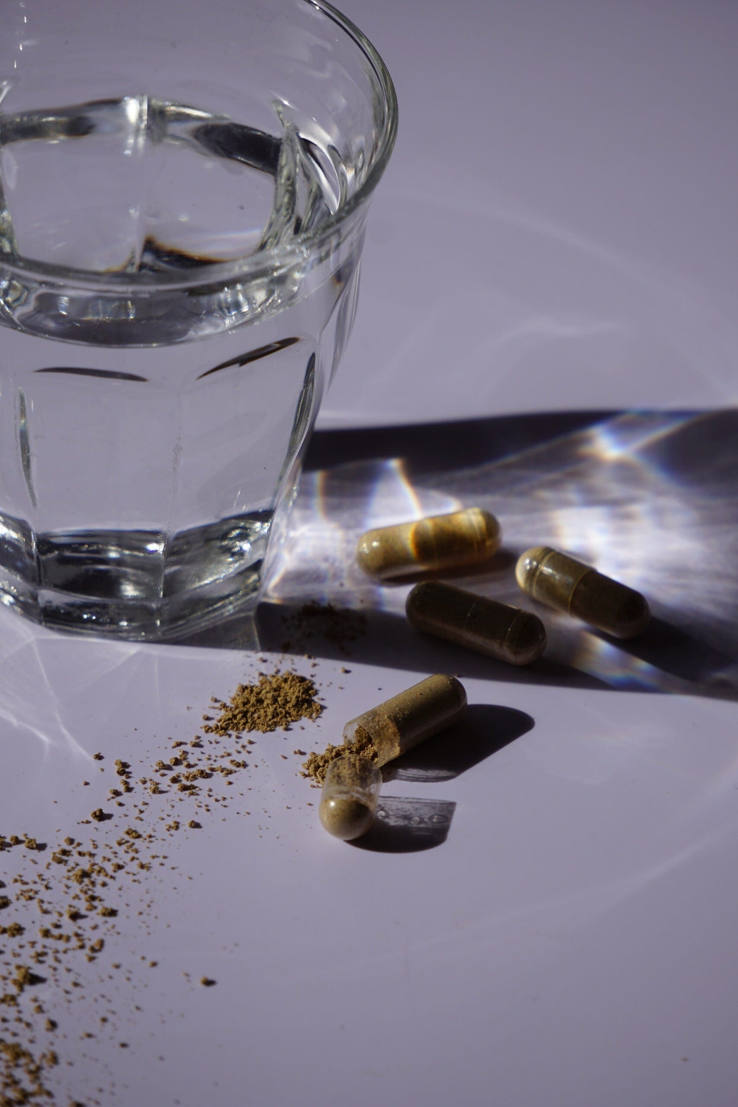 Opened herbal supplement capsules and spilled powder beside a glass of water on a white surface with dramatic reflected light