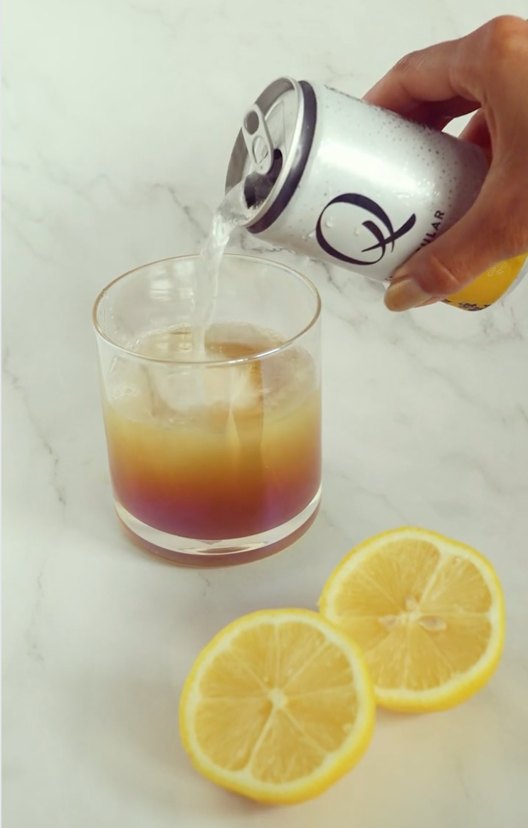 Hand pouring sparkling herbal soda from a can into a layered mocktail in a rocks glass on a marble surface with lemon halves