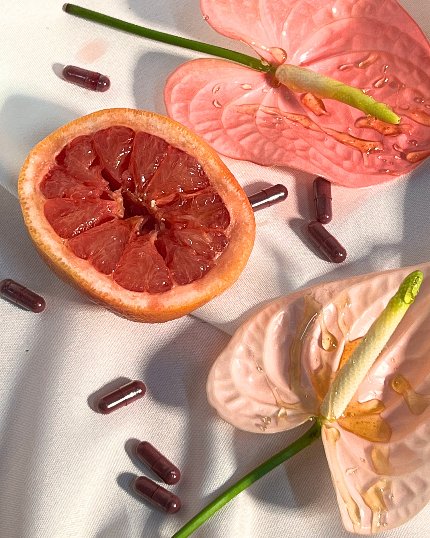 Grapefruit half, pink anthurium flowers, and scattered dark purple supplement capsules on white cloth