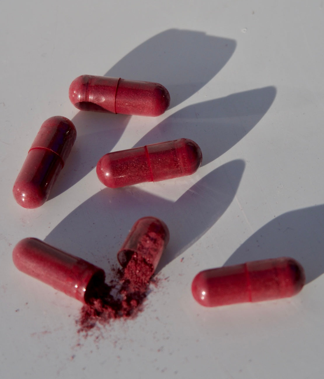 Overhead view of red probiotic capsules showing one opened capsule spilling red powder on a white surface with soft shadows