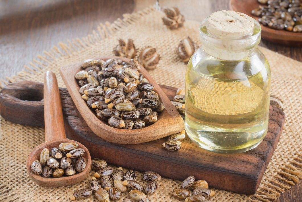 Close-up of glass jar of castor oil and wooden scoops of castor beans showing pale yellow oil and speckled beans on a wooden board