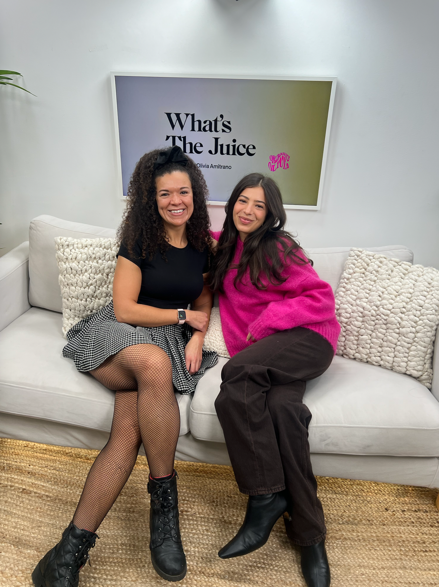 Two women smiling on a couch under a sign reading What’s The Juice, one wearing black and the other a bright pink sweater